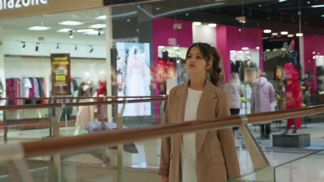 young white woman in mall wandering railing and storefronts, thoughtful gaze scanning displays, casual blazer and jeans, soft interior lights and modern retail atmosphere, slow paced lifestyle moment