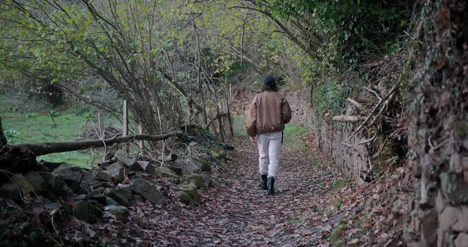 Young woman in a jacket walking on a rural path with a camera. Rear view of a female photographer taking pictures of nature in an autumn forest.