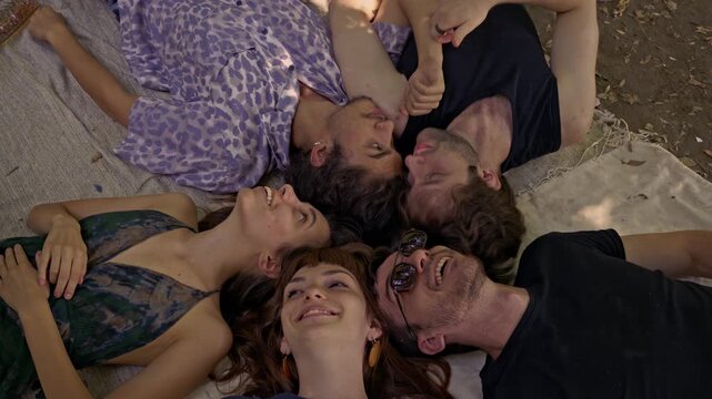 A diverse group of happy friends shares a joyful moment, lying together in a circle on a blanket in nature, laughing and enjoying their time outdoors. - Powered by Adobe