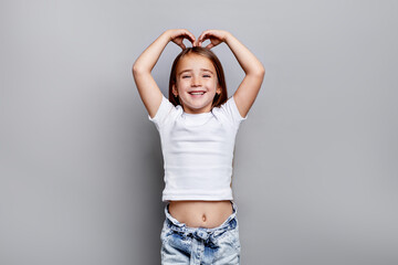 Happy young girl making heart shape with hands above head and smiling in studio, wearing white t-shirt and jeans, joyful portrait against clean grey background