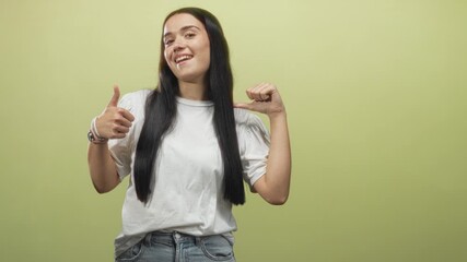 Young woman wearing white tee gives thumbs up with one hand and points thumb to chest while smiling in studio; confidence self belief.