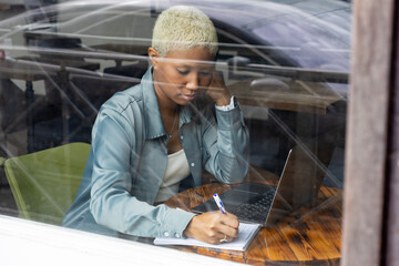 African American woman with short blond hair sits at table and types on laptop. female makes notes in notebook. online education, online work. view through the window of the coffee shop.