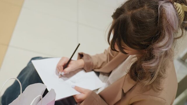 caucasian illustrator sketching on bench with pen and notebook, close overhead view of hands drawing on paper, tiled mall floor, casual jacket and tote beside subject, focused creative atmosphere