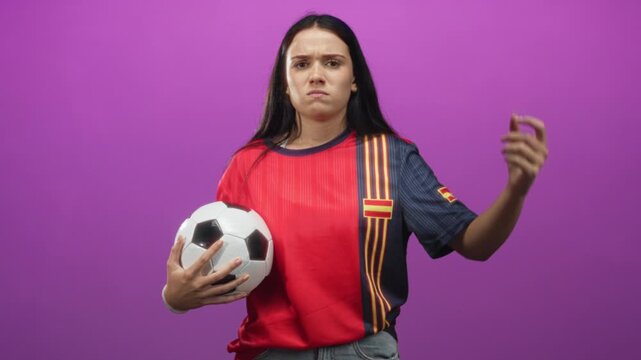 Woman in red spanish jersey holds soccer ball and makes questioning hand gesture in studio; team spirit frustration.