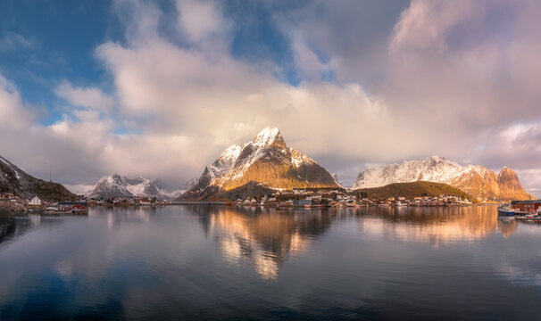 Panoramic aerial view of Hamnoy and Reine villages at sunrise in Lofoten Islands, Norway. Snowy mountains reflected in calm fjord water under cloudy winter sky. Top drone view. Dramatic landscape