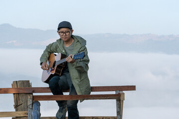 A young traveler plays guitar on a mountaintop balcony, enjoying peaceful morning sunlight and...