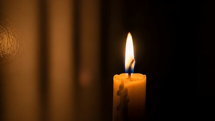 A single burning candle with a warm flame in a dark room with blurred curtains in the background.