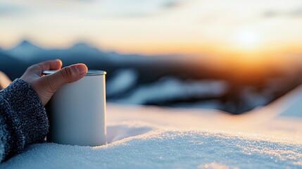 A hand holding a white coffee cup in a snowy landscape, with a beautiful sunset in the background.