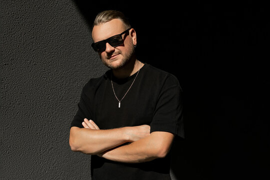 Portrait of stylish beard man in sunglasses standing against a gray dark background with arms crossed