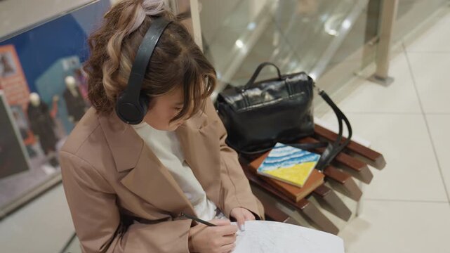 woman headphones sketching lyrics notebook on tiled floor bench near retail displays, stack of books and black bag at side, intimate closeup of handwriting and contemplative expression