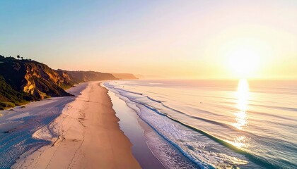 Aerial view of a beach at sunset with golden light reflecting on the water and sand. Cliffs and vegetation are visible along the coastline.
