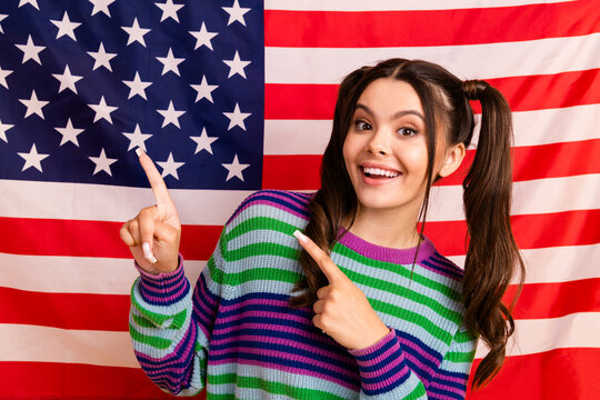 Young woman with a bright smile points to the american flag while wearing a colorful striped sweater for a cheerful patriotic lifestyle image - Powered by Adobe