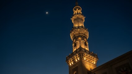 A lit tower with ornate details stands against a dark blue night sky with a small moon.