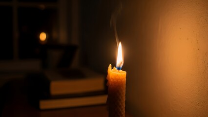 A single lit candle standing on a table next to stacked books in a dark room with a warm glow on the wall.