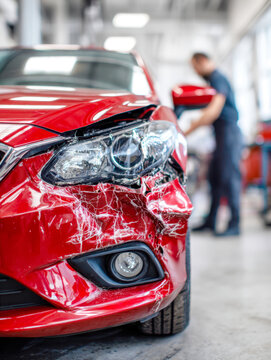 Red car with severe front-end damage and cracked headlight in an auto repair shop with technician inspecting the vehicle in the background