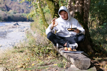 Young man in hoodie making okay gesture while eating from container by tree near river