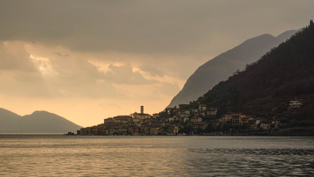 An Italian village perches on the edge of the mountains overlooking a lake, with a central church standing tall amid warm yellow-orange light from the setting sun.
