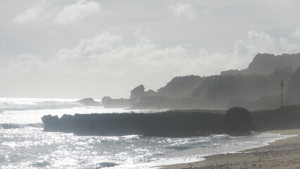 A foggy afternoon shrouds the coastline, where gentle waves glitter subtly under muted light, creating a serene and mysterious seascape.