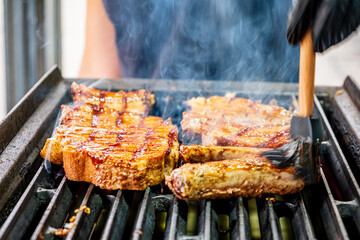 Steaks sizzle on a grill, coated in a rich sauce, releasing savory aromas on a sunny day