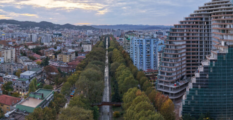 Aerial view of Tirana featuring the Lana River, dense residential districts, and a modern high-rise under construction.