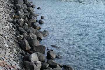Close-up view of a rocky embankment where dark stones meet calm blue water, creating a natural coastal or lakeside texture.
