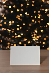 Festive greeting postcard mockup, blank paper card on table, blurred Christmas golden shining lights bokeh on background