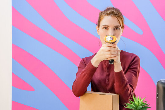 Woman aiming barcode scanner at nose with package on table against colorful stripes