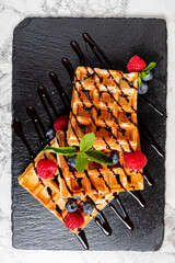 Two golden waffles are beautifully decorated with berries and chocolate on a sleek black slate