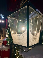 Decorative vintage electric lantern with snow inside, illuminated by warm light, surrounded by festive holiday decorations, creating a cozy winter night atmosphere. Vertical background
