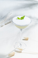 A beautifully crafted cocktail with frothy foam and mint leaves sits elegantly on a light surface