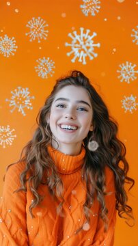 A young woman with long brown hair and blue eyes is smiling towards the camera. She wears a warm orange sweater. In the background, there's an orange wall with white snowflake decals on it.