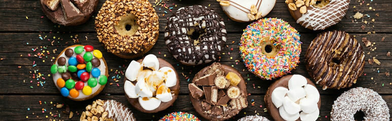 Fototapeta premium Wide Image of Four donuts featuring chocolate sprinkles, white drizzle, and white icing arranged on a beige napkin resting on a wooden table