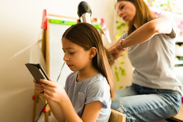 Mother drying daughter's hair while child is using a tablet, morning routine with digital entertainment