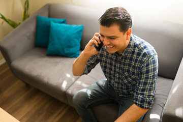 Fototapeta premium Happy man having a phone call on a couch at home, smiling while talking and enjoying wireless communication