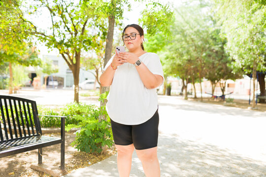 Active overweight woman wearing glasses, a white t-shirt, and shorts, standing in a park, focused on her smartphone