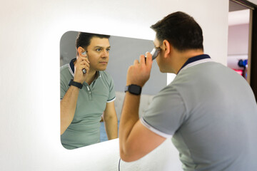 Adult man trimming ear hair using electric razor in front of a mirror in a bathroom, focusing on personal grooming and self-care