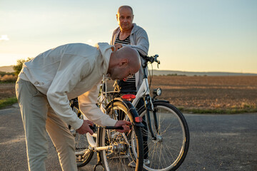 Two men with bicycles on rural road sunset, one fixing tire while other watches, teamwork and...