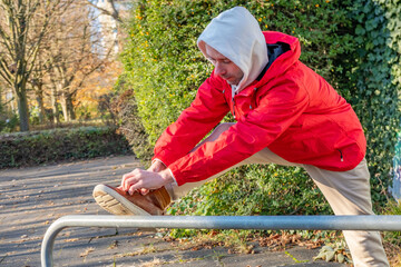 young man in red jacket stretching leg on park railing, warmup before run, solo training in cold...