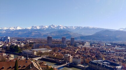 A stunning, wide view of Grenoble's urban sprawl set against the dramatic, snow-capped peaks of the Belledonne mountain range. The Isère River and the Three Towers stand out.