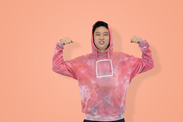 Strong and smiling young Asian man in a pink hoodie flexing both biceps, symbolizing health, confidence, youthful power, and determination in pink studio background