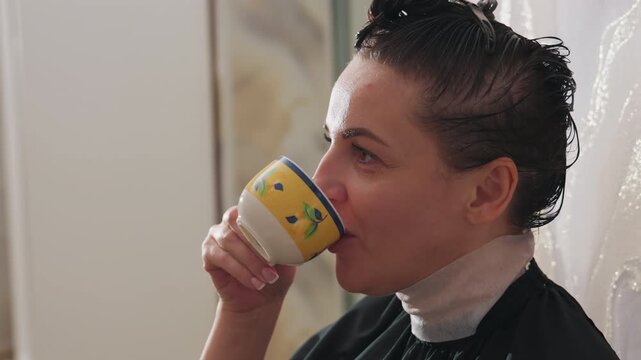 white woman salon client sipping tea, wet hair under cape during hair treatment, calm introspective expression, ceramic mug closeup, soft natural light, neutral interior, stylist prep implied, candid