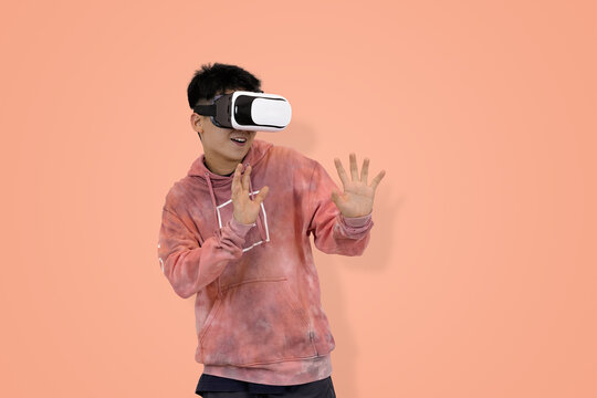 Shocked young Asian man in hoodie wearing a virtual reality headset with his hands raised, reacting in surprise to an immersive gaming or metaverse experience in studio background
