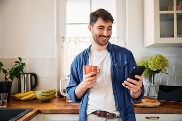 Young smiling man using mobile phone while drinking morning coffee at home