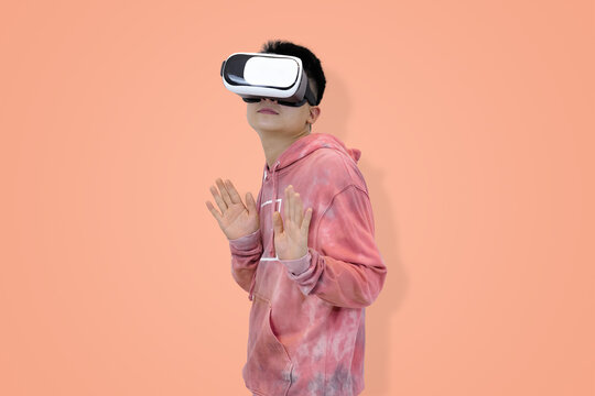 Shocked young Asian man in hoodie wearing a virtual reality headset with his hands raised, reacting in surprise to an immersive gaming or metaverse experience in studio background
