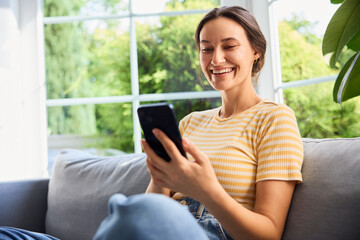 Fototapeta premium Cheerful young woman using mobile phone sitting on sofa at home