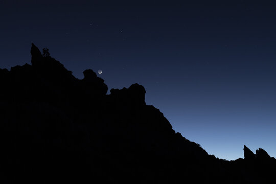 View of a dark, jagged silhouette of a rocky landscape under a gradient of deep blue and black sky with a faint crescent moon, Tenerife, Canary Islands, Spain.