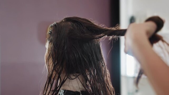 caucasian woman having hair sectioned and brushed for blowout, hairdresser lifting sections with brush, salon mirror glow and preparation for styling, brisk confident salon energy and professional