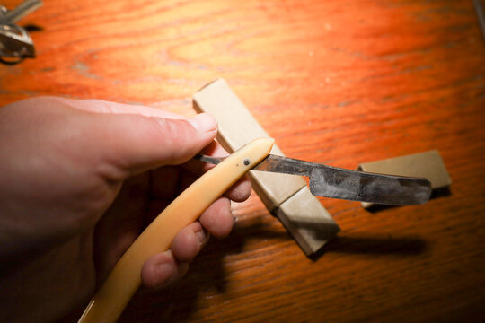 A person's hand holds a vintage straight razor with a bone or faux-bone handle and an aged metal blade over a wooden surface. The razor is open and positioned diagonally above a light beige cardboard 
