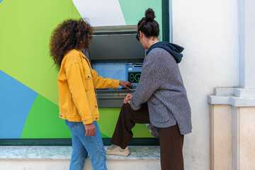 Young women making financial transaction at an automated teller machine, partner assisting outdoors