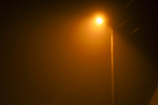 A bright orange streetlight on a tall pole pierces through thick fog or haze on a dark night. The light creates a halo, giving the scene a mystical and enigmatic appearance.